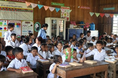 Schoolchildren in white uniforms hold up new books at their wooden desks in a decorated classroom